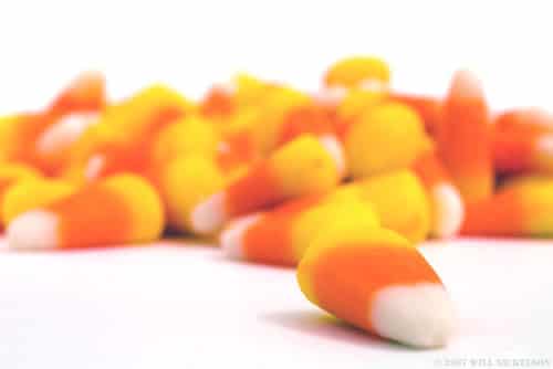 A closeup of candy corn, with one kernel in the foreground and an out-of-focus pile in the background, against a white backdrop.