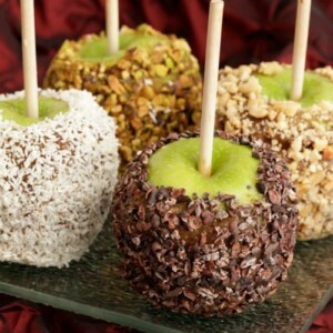 Four caramel green apples, with different coatings, including nuts, coconut, and cacao nibs.