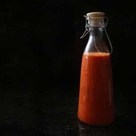 A tall jar of red hot sauce, against a stark black background.