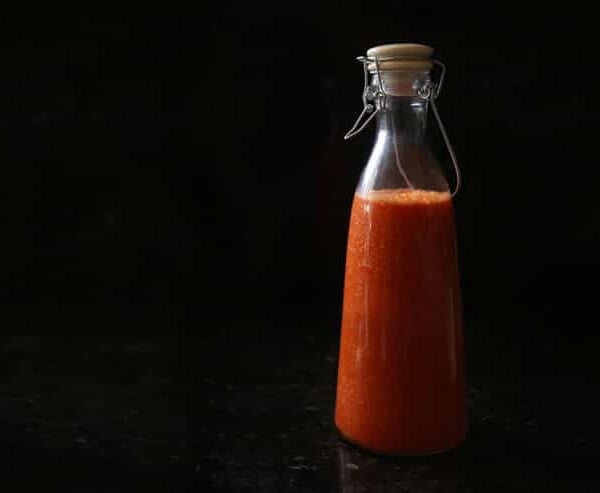 A tall jar of red hot sauce, against a stark black background.